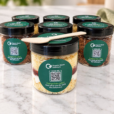 Branded cake jars with QR code sticker. Inside the cake jars are layered with sponge, jam and buttercream, with a spoon for serving. 