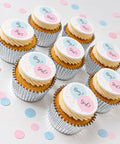 several gender reveal cupcakes shown on a white table with pastel confetti. Each cupcake has an edible topper saying boy or girl. Secret coloured filling inside. 