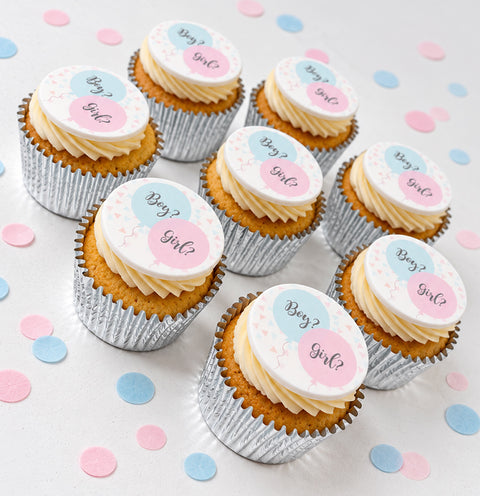several gender reveal cupcakes shown on a white table with pastel confetti. Each cupcake has an edible topper saying boy or girl. Secret coloured filling inside. 