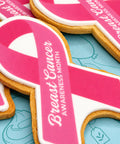 Pink and white breast cancer awareness ribbon-shaped biscuit with white icing text reading 'Breast Cancer Awareness Month,'.