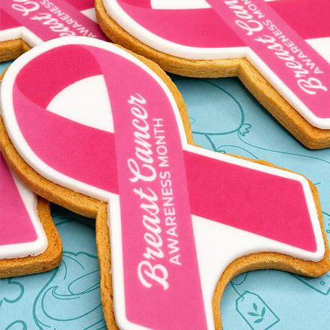 Pink and white breast cancer awareness ribbon-shaped biscuit with white icing text reading 'Breast Cancer Awareness Month,'.