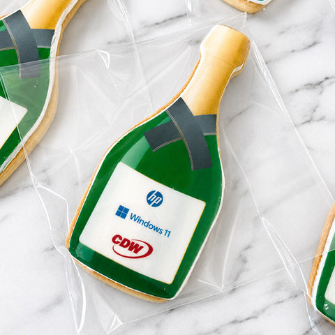 A close-up of a shaped sugar cookie decorated to look like a green champagne bottle with gold foil, a white label featuring HP, Windows 11, and CDW logos, and dark gray ribbon accents, shown wrapped in clear cellophane on a white marble surface.