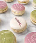 A styled flat-lay of printed macarons on a white marble surface. The macarons feature pastel pink, green, and cream shells with  illustrations and company logo.