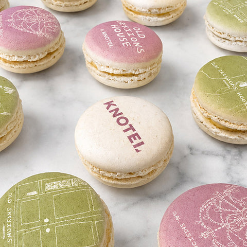 A styled flat-lay of printed macarons on a white marble surface. The macarons feature pastel pink, green, and cream shells with  illustrations and company logo.
