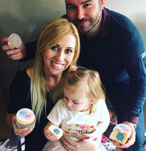 A family doing a gender reveal with cupcakes. A mother and father are shown with a toddler, all holding a cupcake.