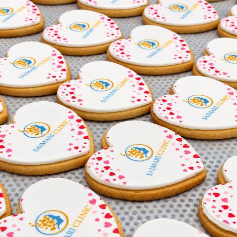 Heart-shaped branded biscuit for Valentines day decorated with white icing, pink heart confetti, and a blue and gold logo that reads 'SAMAHARI CLINIC'.