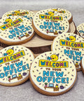 Round sugar cookies decorated with a fun 'Welcome to Your New Office!' design, arranged on a rustic wooden slice and a light grey surface.