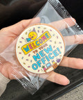 A hand holds a round white cookie decorated with a colorful 'Welcome to your new office!' design, sealed inside a clear plastic bag.