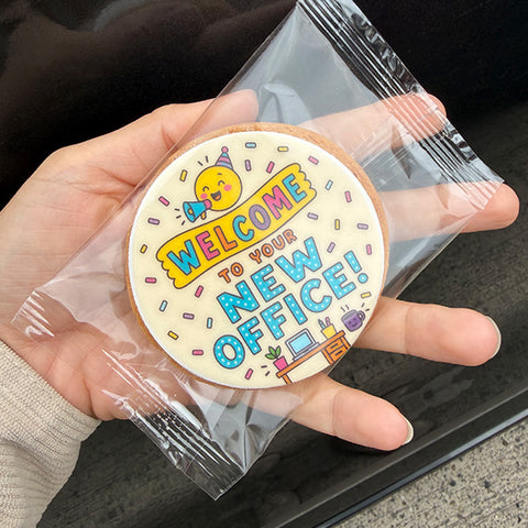 A hand holds a round white cookie decorated with a colorful 'Welcome to your new office!' design, sealed inside a clear plastic bag.
