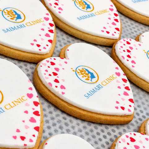 Heart-shaped sugar cookies with white icing, decorated with pink and red heart confetti, a blue and gold clinic logo, and  'SAIMARI CLINIC' logo.