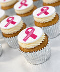 A close-up of several frosted vegan cupcake, each topped with a white round topper featuring a pink ribbon and Breast cancer awareness month.