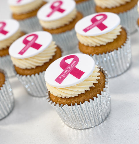 A close-up of several frosted vegan cupcake, each topped with a white round topper featuring a pink ribbon and Breast cancer awareness month.