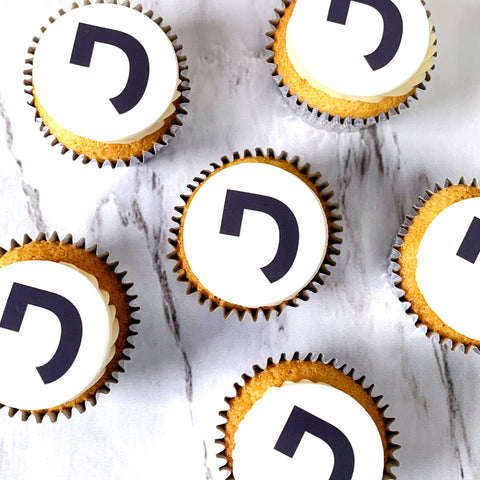 Logo branded cupcakes baked in silver cased with a buttercream swirl. Each cupcake is topped with a n edible logo print and they are displayed on a marble worktop. 